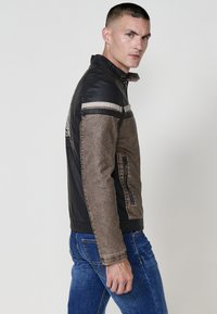 Uomo con capelli corti che indossa una giacca di pelle nera e marrone e jeans blu, che guarda oltre la spalla su uno sfondo chiaro e semplice.