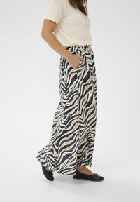 Kvinde iført brede zebra-stribede bukser, beige t-shirt, sorte ballerinaer, med den ene hånd i lommen, stående på hvid baggrund.