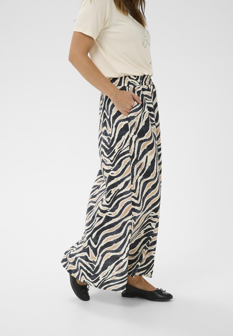 Kvinde iført brede zebra-stribede bukser, beige t-shirt, sorte ballerinaer, med den ene hånd i lommen, stående på hvid baggrund.