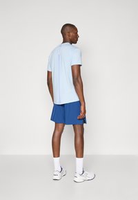 Lysblå kortærmet skjorte med en vertikal logo detalje på ryggen, parret med mørkeblå shorts og hvide sportssko.