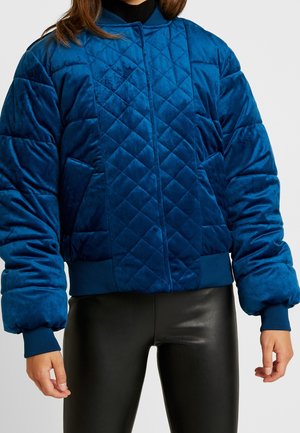 Kvinde iført en quiltet, polstret blå fløjls bomberjakke med ribbede manchetter, kant og krave, kombineret med sorte læderbukser.