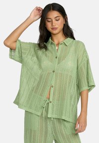 Camicetta verde chiaro, a maglia aperta, con maniche corte e chiusura a bottoni, dal fit rilassato. Abbinata a pantaloni coordinati, con texture e colore simili.