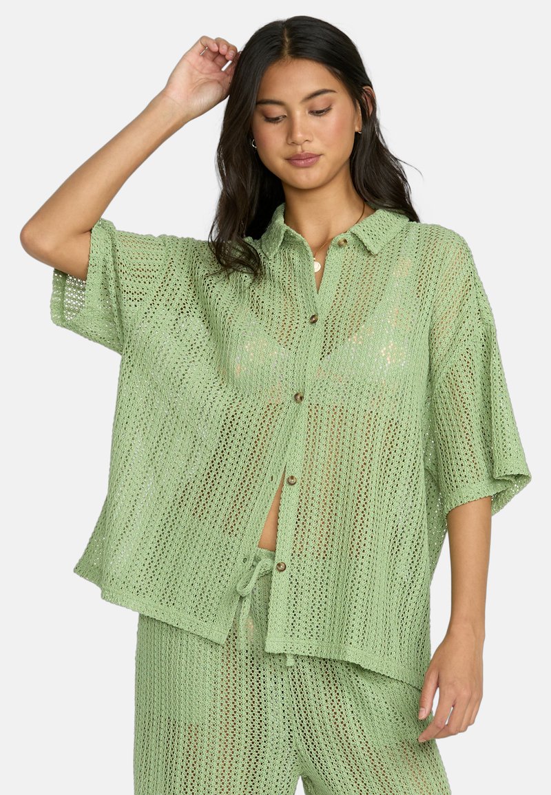 Camicetta verde chiaro, a maglia aperta, con maniche corte e chiusura a bottoni, dal fit rilassato. Abbinata a pantaloni coordinati, con texture e colore simili.