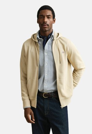 Man wearing beige zip-up hoodie over striped button-up shirt, dark neck scarf, braided belt, and dark jeans, standing with one hand behind back.