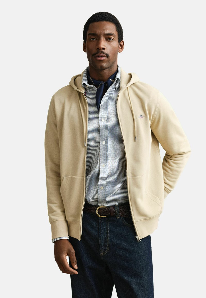 Uomo che indossa una felpa beige con zip sopra una camicia a righe con bottoni, sciarpa scura intorno al collo, cintura intrecciata e jeans scuri, in piedi con una mano dietro la schiena.