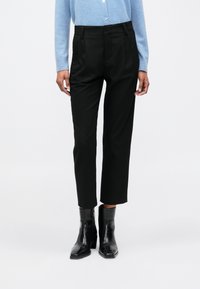 Femeie îmbrăcată în pantaloni negri scurți, cizme negre până la gleznă și o bluză albastră deschis cu mânecă lungă și nasturi, stând pe un fundal alb.