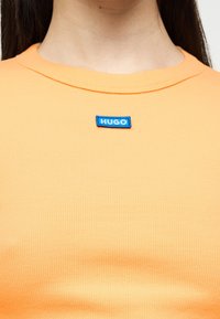 Gros plan sur une personne portant un t-shirt orange côtelé avec un petit patch logo rectangulaire bleu "HUGO" au centre de la poitrine.