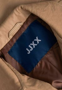 Rudos odos striukės vidus su tamsiai mėlynu etiketė „JJXX“. Turtingas lygus paviršius ir kontrastinga ruda pamušalo spalva.