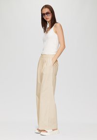 Witte geribde tanktop gecombineerd met beige, wijde broek met een gestructureerde afwerking. Accessoires omvatten oversized zonnebrillen en witte sandalen.
