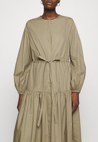 Donna che indossa un abito lungo con maniche, di colore beige, con scollatura rotonda, vita cintrata legata con un fiocco e dettagli di gonna arricciata.