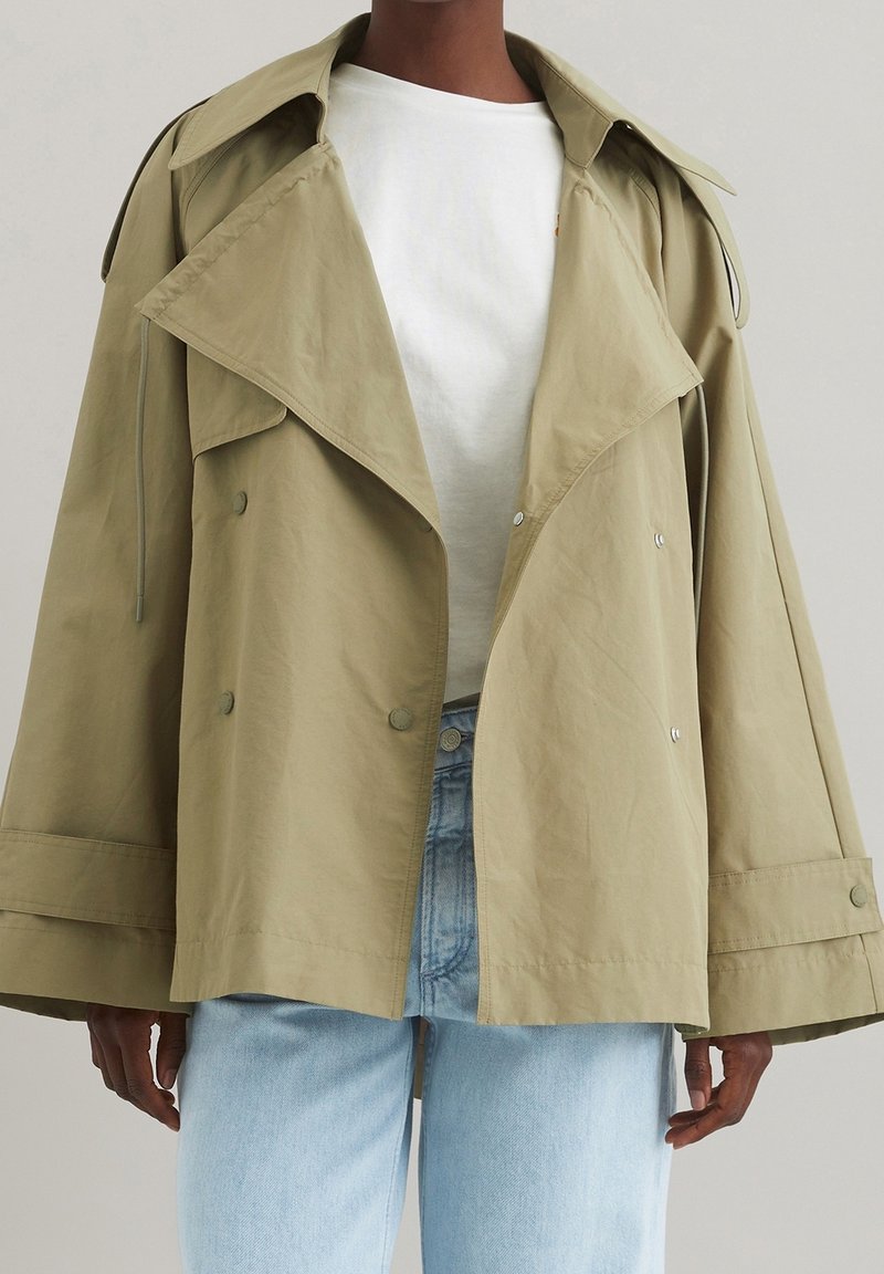 Beige oversized trenchcoat med bred krave, knaplukninger og sidelukninger. Båret over en hvid skjorte og lyseblå jeans.