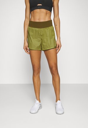 Slank person iført olivengrønne sportsshorts med bred talje, sort sports-bh og hvide løbesko, stående mod en hvid baggrund.