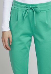 Türkise spordipükse, millel on paelaga vöökoht, ees on pleidid ja sile tekstuur. Üksikasjalik pilt käest, mis toetub jalale.