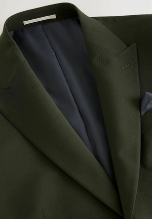 Tamnozeleni blejzer s velikim reverima i glatkim tamnoplavi podstavu. Sadrži diskretno šivanje i lijevi džep na prsima.