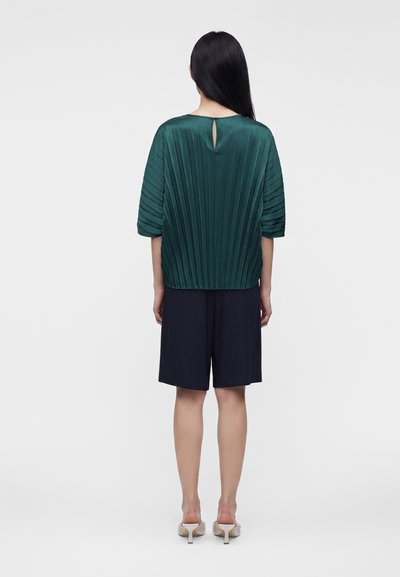 Femme aux longs cheveux foncés portant un haut plissé vert et un short noir jusqu’aux genoux, debout de dos sur un fond blanc.