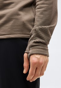 Közeli kép egy kézről, amely barna, hosszú ujjú, texturált felsőt visel hüvelykujj nyílással, fekete nadrággal világos háttér előtt.