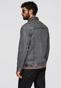 Giacca in denim grigia con taglio classico, caratterizzata da una chiusura frontale con bottoni, colletto a punta e dettagli cuciti. Indossata sopra una maglia dai toni neutri.