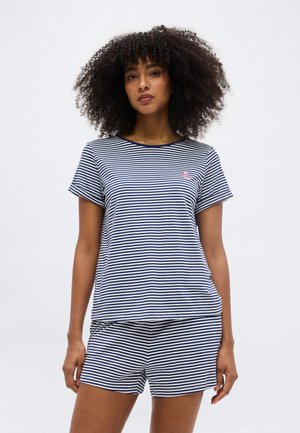 Göndör hajú nő sötétkék és fehér csíkos rövid ujjú felsőt és hozzá illő rövidnadrágot visel, egy egyszínű háttér előtt áll.
