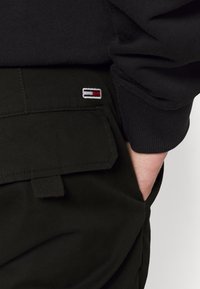 Zwarte pantalon van een gestructureerde stof, met een klein geborduurd logo met rode, witte en blauwe accenten op de achterzak.