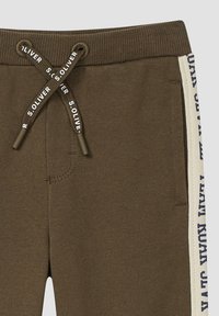 Braune Sweatpants mit geripptem Bund, ausgestattet mit einem Kordelzug und seitlichen Streifen mit blauer Schrift auf cremefarbenem Hintergrund.