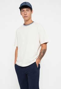 Short-sleeve cream t-shirt with a navy ribbed collar, paired with navy pants. A tattoo is visible on the left arm.