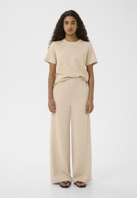 Beige Baumwoll-T-Shirt mit kurzen Ärmeln, kombiniert mit weit geschnittenen passenden Hosen. Glatte Textur, lässige Passform und minimale Designmerkmale. Flip-Flops sichtbar.