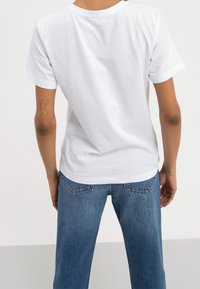 T-shirt blanc à manches courtes avec un col rond, design sobre, texture douce, associé à un pantalon en jean bleu clair à jambes droites et un tatouage visible sur le bras.