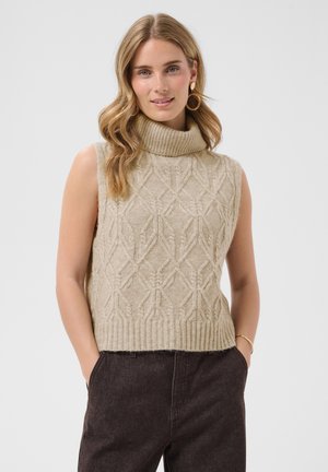 Ung kvinde med bølget blondt hår iført beige ærmeløs kabelstrikket højhalset sweater og mørke bukser, står med hænderne i lommerne.