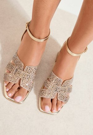 Füße in goldenen, offenen Sandalen mit Riemen am Knöchel, verziert mit Strasssteinen, die auf einem hellen Boden stehen.