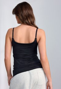 Débardeur noir en tissu lisse et extensible avec fines bretelles et encolure arrondie, porté avec un pantalon de survêtement gris clair.