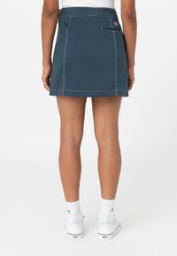 Modré šortky s bílým šitím, zadní kapsou na zip a plochým pasem. Nošeny s bílými ponožkami a teniskami, zobrazené z zadní strany.