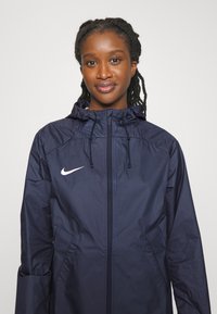 Giacca a vento blu navy con cappuccio, chiusura con zip, cordini e logo Nike bianco sul lato sinistro del petto. Presenta tasche laterali.