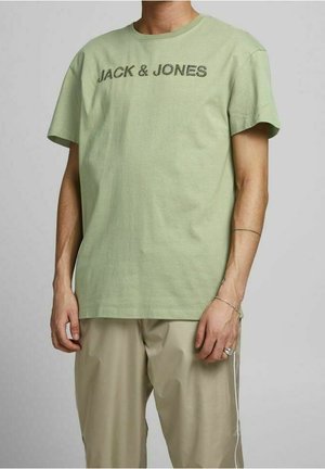 Mann trägt ein hellgrünes "JACK & JONES"-T-Shirt und beige Hose, mit minimalem Schmuck am linken Handgelenk und an den Fingern.