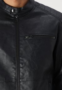Giacca in pelle nera con colletto alto, dotata di chiusura frontale con zip e tasca sul petto con zip. Texture liscia e design attillato.