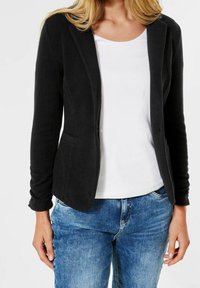 Musta istuva bleiseri pehmeästä kankaasta, jossa on yhden napin suljenta, kapeat läpät, kaksi taskua ja paritettu valkoisen paidan ja sinisten farkkujen kanssa.