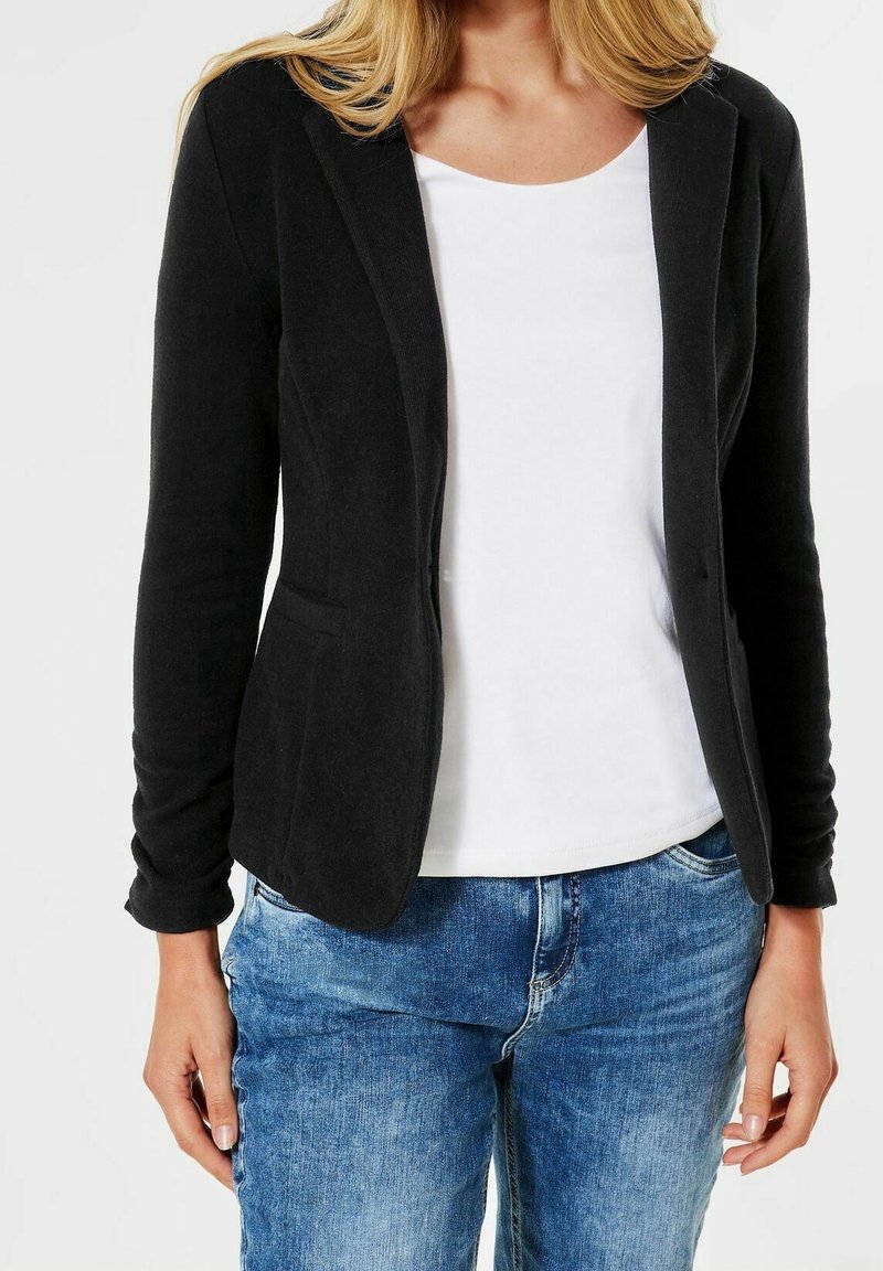 Musta istuva bleiseri pehmeästä kankaasta, jossa on yhden napin suljenta, kapeat läpät, kaksi taskua ja paritettu valkoisen paidan ja sinisten farkkujen kanssa.