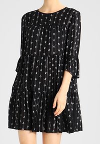 Schwarzes Kleid mit rundem Ausschnitt, dreiviertellangen Ärmeln und einem gestuften Saum. Verfügt über silberne Diamantmuster und eine fließende Stofftextur.