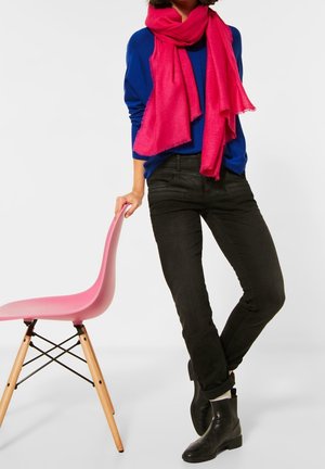 Blauwe trui met lange mouwen, roze sjaal met franjes, zwarte slim-fit jeans en zwarte enkellaarsjes. Roze stoel met houten poten op de achtergrond.