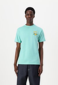 T-shirt de algodão azul-turquesa de mangas curtas, apresentando um gráfico floral amarelo e verde no lado esquerdo do peito. Decote redondo e corte descontraído.