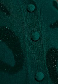 Gros plan sur un tissu tricoté vert foncé avec trois boutons texturés et des taches noires duveteuses et scintillantes.
