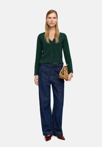 Zielony sweter z dzianiny z ozdobnymi guzikami, w połączeniu z luźnymi niebieskimi dżinsami i torebką w panterkę. Brązowe buty na szpilce.