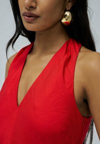 Vestido rojo sin mangas con un escote en V profundo, hecho de una tela suave. Cuenta con un cierre lateral y un diseño drapeado. Pendientes circulares dorados que añaden contraste.