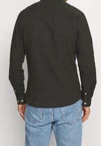 Camicia verde scuro a maniche lunghe realizzata in tessuto strutturato, con polsini abbottonati e un motivo sul dietro. Abbinata a jeans azzurri.