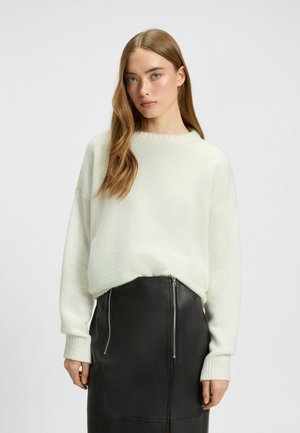 Maglione oversized color crema con collo e polsini a coste, abbinato a una gonna in pelle nera con dettagli con zip. Tessuto morbido.