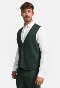 Mørkegrønn herringbone vest laget av teksturert stoff, med dyp V-hals, fem knapper og en frontlomme, båret over en hvit skjorte.