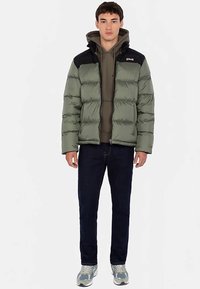 Doudoune vert olive avec accents noirs aux épaules, dotée d'une fermeture éclair frontale, d'une texture matelassée et d'une silhouette ajustée.