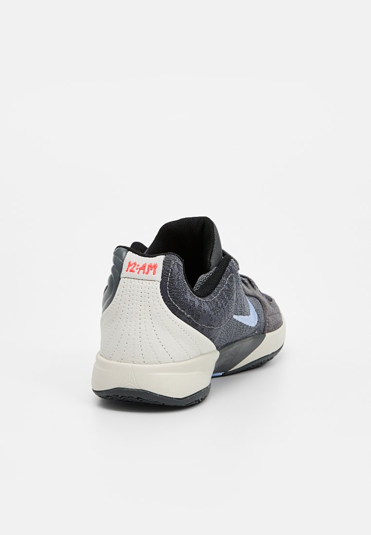 Zapatilla deportiva en negro y gris con parte superior texturizada, talón blanco y logo Swoosh azul. Presenta un exclusivo texto "12 AM" en el talón.