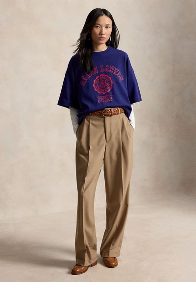 Γυναίκα που φοράει μπλε Ralph Lauren γραφική μπλούζα πάνω από λευκές μακριές μανσέτες, μπεζ πιετά παντελόνια με πλεκτή ζώνη και καφέ μοκασίνια.