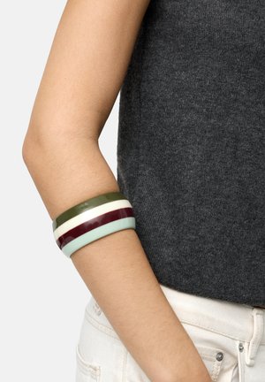 Ramię noszące szeroką, gładką bransoletkę typu bangle z poziomymi paskami w kolorach zielonym, białym, bordowym i jasnoniebieskim, na ciemnoszarym topie.