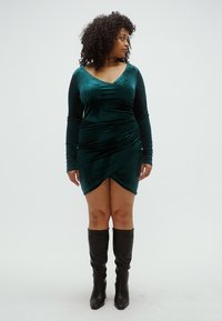 Βελούδινο πράσινο φόρεμα με μακριά μανίκια, σχέδιο τύπου γύρισμα και ρυτιδωμένες λεπτομέρειες. Συνδυασμένο με μαύρες μπότες μέχρι το γόνατο. Λείο ύφασμα.